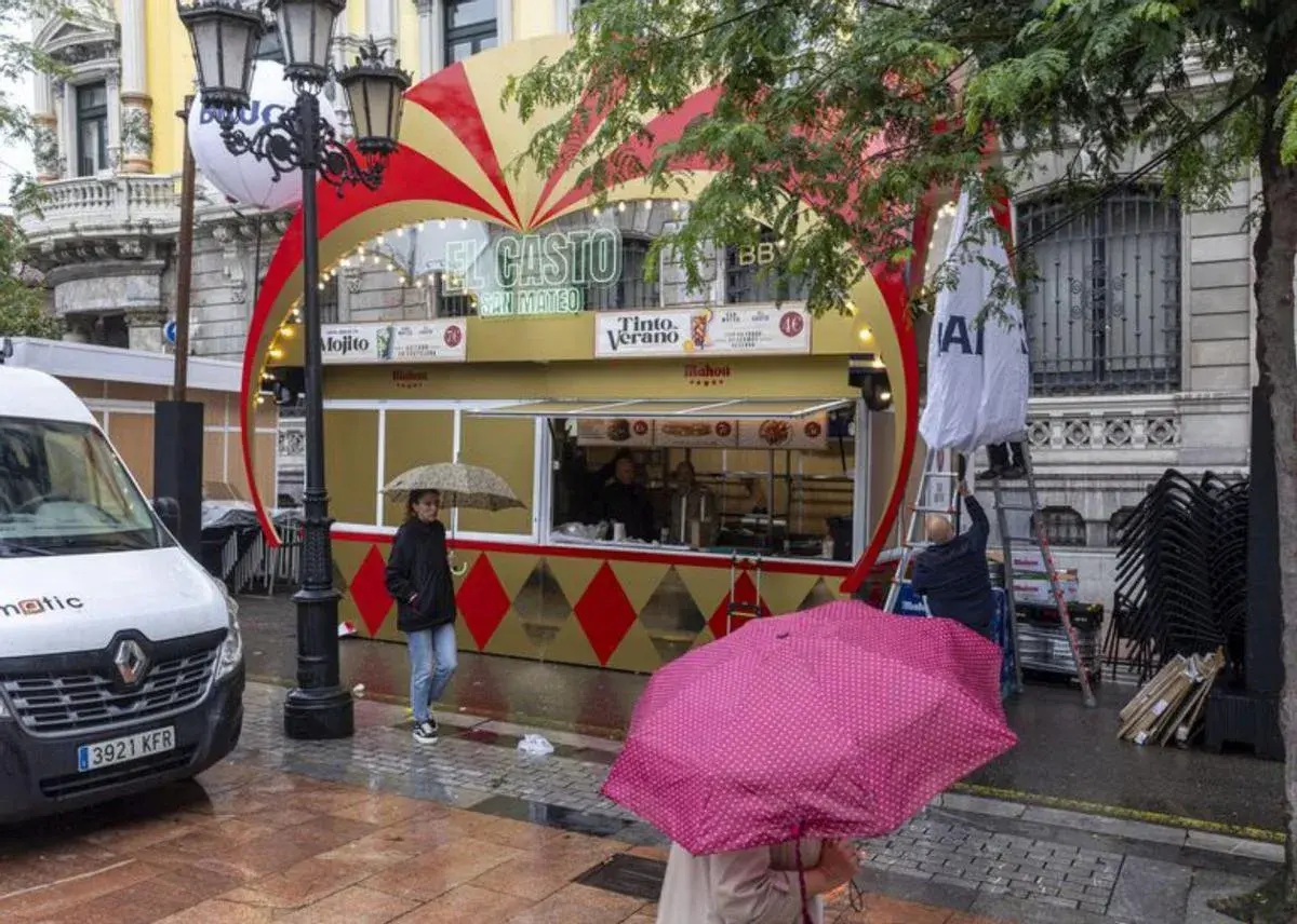 Preparativos para las fiestas de San Mateo en Oviedo. Un puesto de comida y bebida con decoración festiva, gente con paraguas y un furgón blanco.