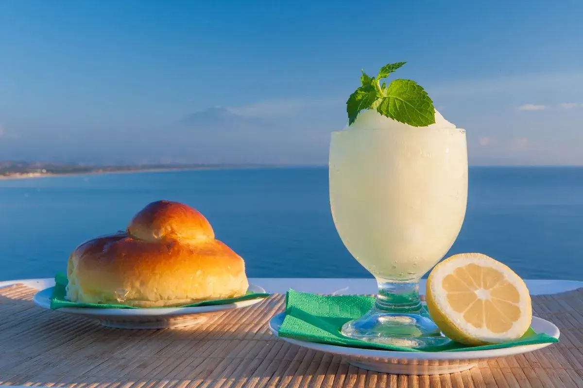 Colazione siciliana con brioche e granita al limone, con il mare e l'Etna sullo sfondo.