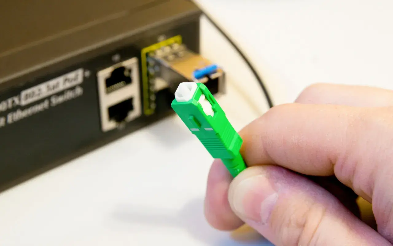 Dłoń trzyma zieloną wtyczkę światłowodową. Obok widać przełącznik Ethernet. Tak sprawdzisz, czy jest światłowód.