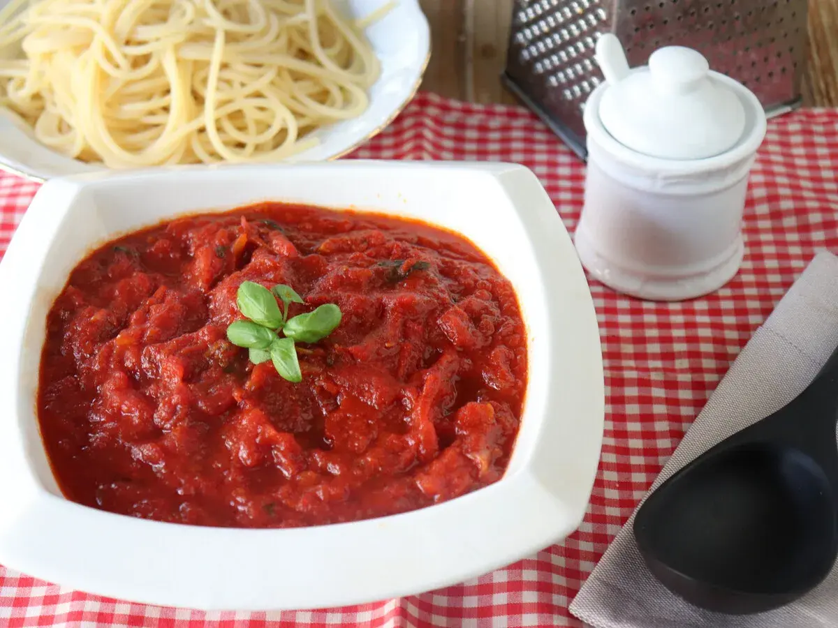 Un tazón blanco lleno de salsa marinara casera, decorada con albahaca fresca, junto a espaguetis cocidos.