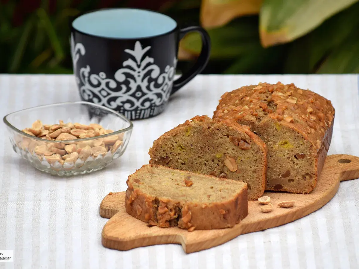 Un delicioso bizcocho de plátano, cubierto de frutos secos, acompañado de cacahuetes y una taza.