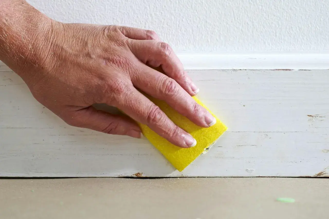 Hand med gul svamp slipar en vitmålad list. Förberedelser för att måla tunt.