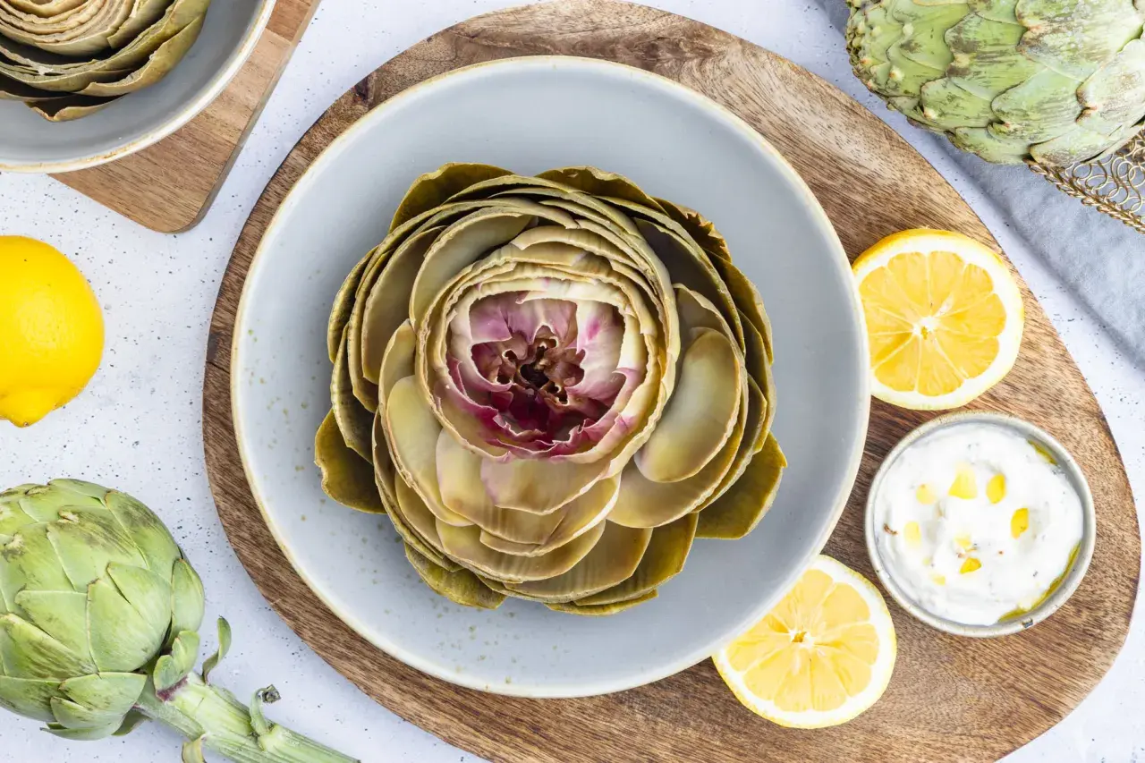 Ein gekochter Artischocke auf einem Teller, bereit für ein leckeres Artischocken Rezept. Dazu Zitronenspalten und eine Dip-Schale.