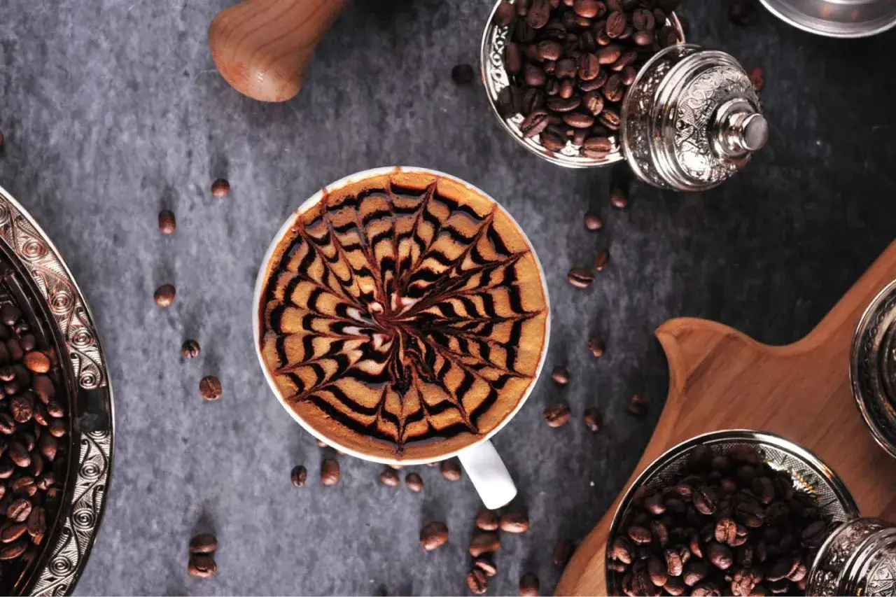Kawa mocha z artystycznym wzorem na piance. Obok ziarna kawy w ozdobnych naczyniach.