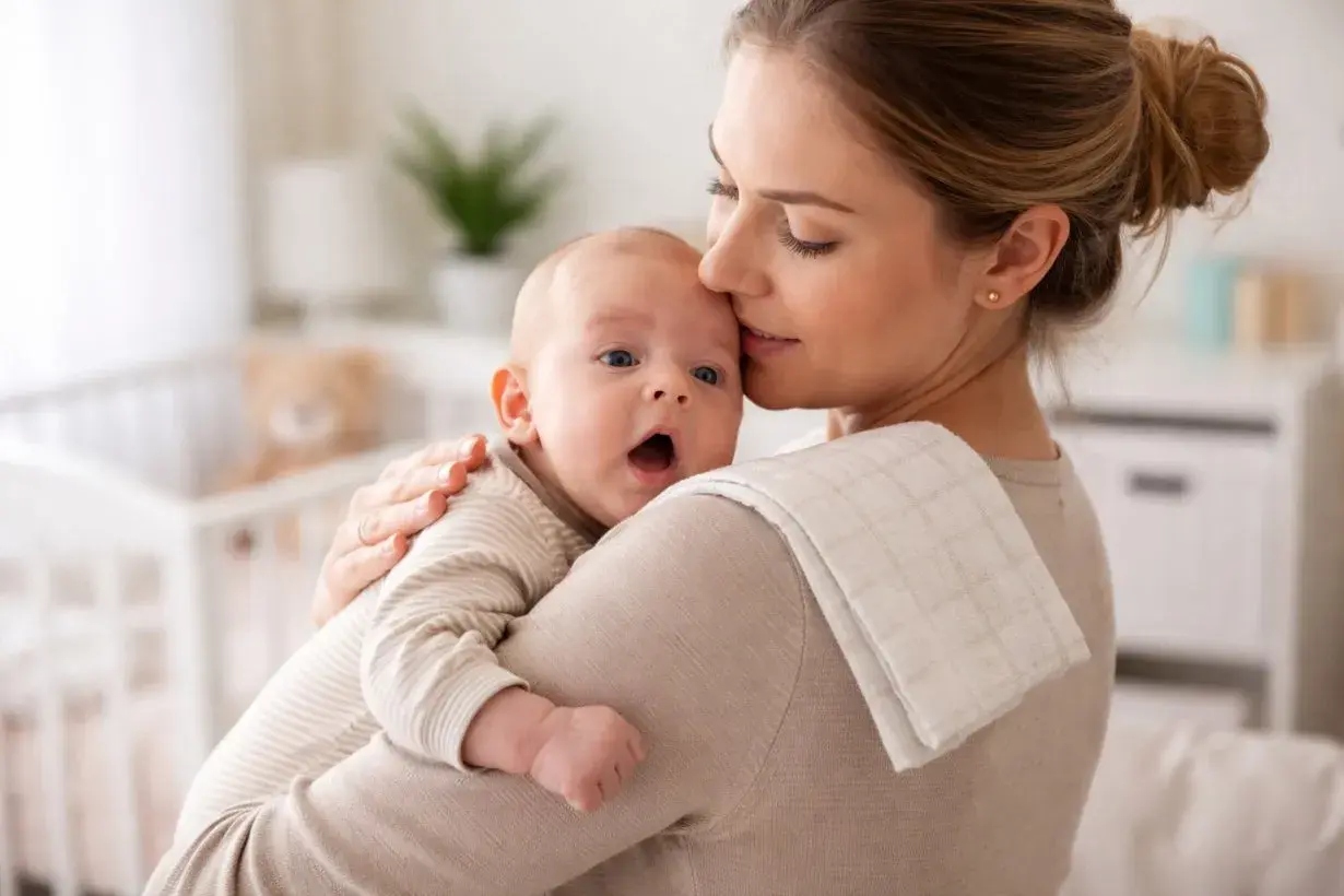 Mamma abbraccia il suo bimbo, che ha un piccolo singhiozzo dopo poppata. La culla e un orsetto sullo sfondo.