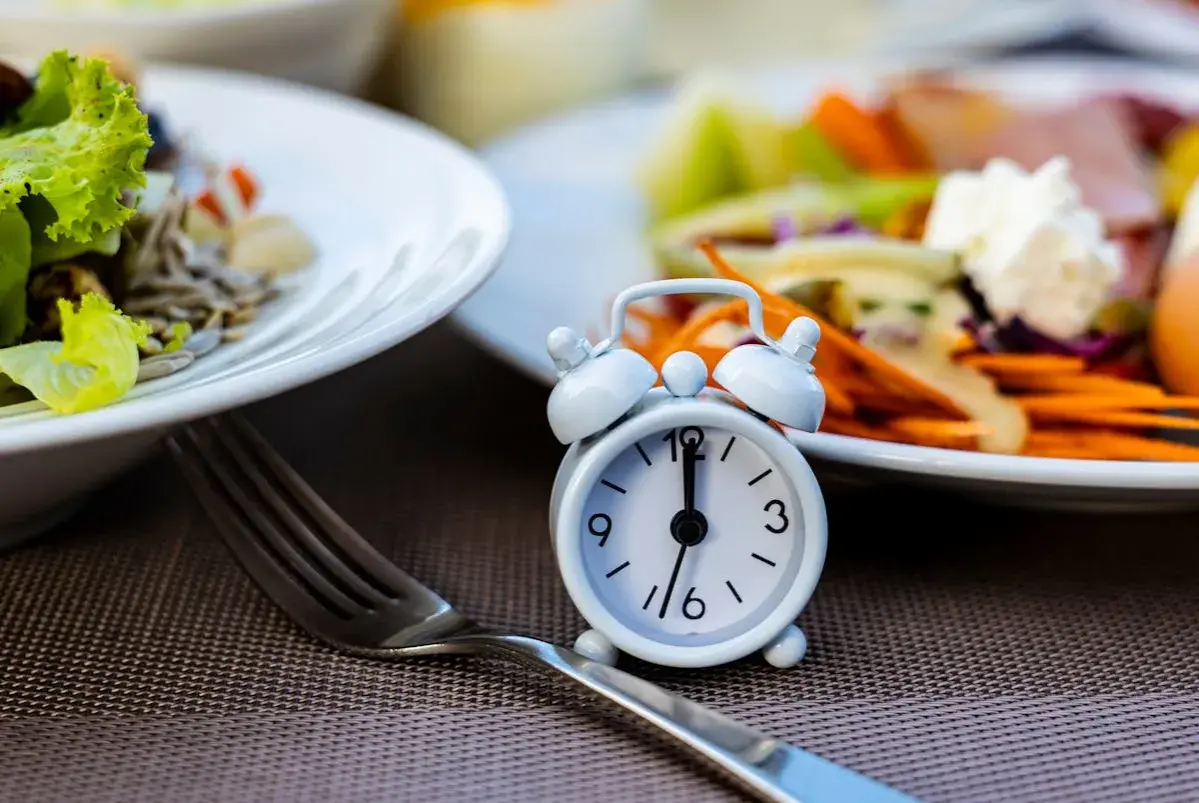 Réveil miniature sur une table, symbolisant le jeûne intermittent 12h. Des assiettes de salade et de légumes sont floues en arrière-plan.
