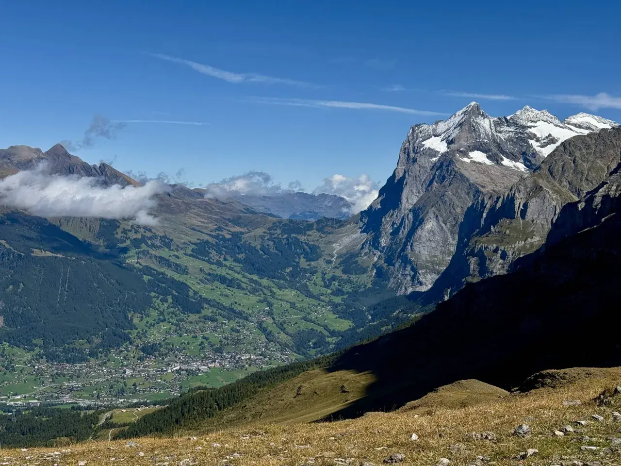 Panoramic view of a valley village nestled among green hills, with dramatic, snow-capped peaks of the Swiss Alps in the background. This scene exemplifies the best hikes in Swiss Alps.