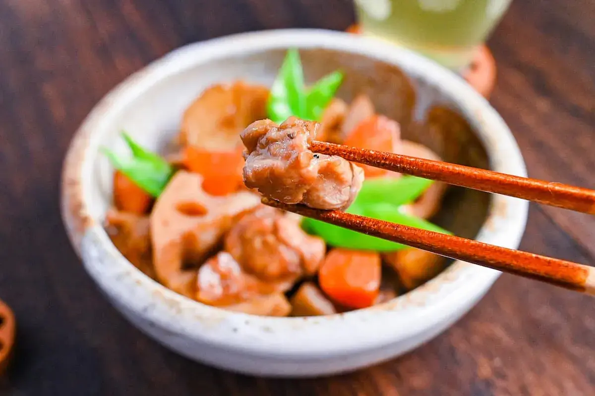 A close-up of a bowl of chikuzenni recipe, with chopsticks lifting a piece of chicken. The dish contains chicken, lotus root, carrots, and green beans.