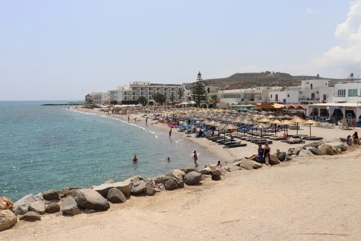 Słoneczna plaża na wyspie Kos z białymi hotelami, parasolami i turystami kąpiącymi się w morzu.