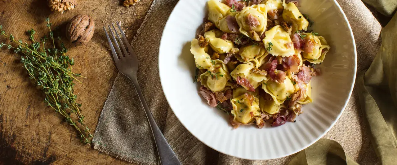 En skål med tortellini prosciutto, valnötter och timjan. En gaffel ligger bredvid på en rustik träyta.
