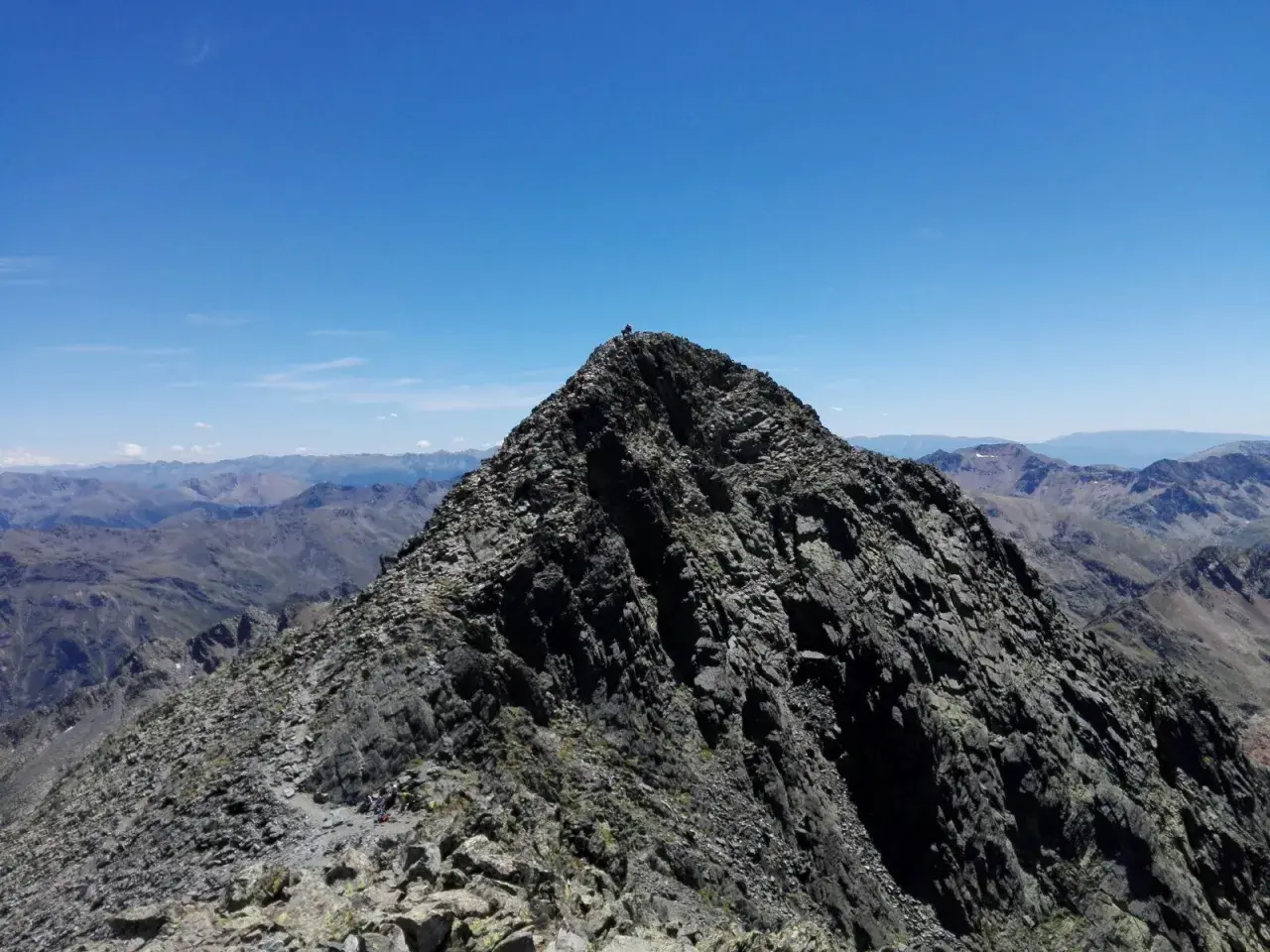 Cima rocosa de montaña con excursionistas y vista panorámica de picos bajo un cielo azul.
