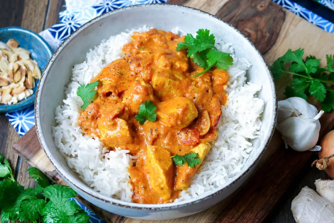 Schüssel mit duftendem Reis und cremigem Chicken Tikka, garniert mit frischem Koriander. Daneben Mandelsplitter, Knoblauch und Ingwer.