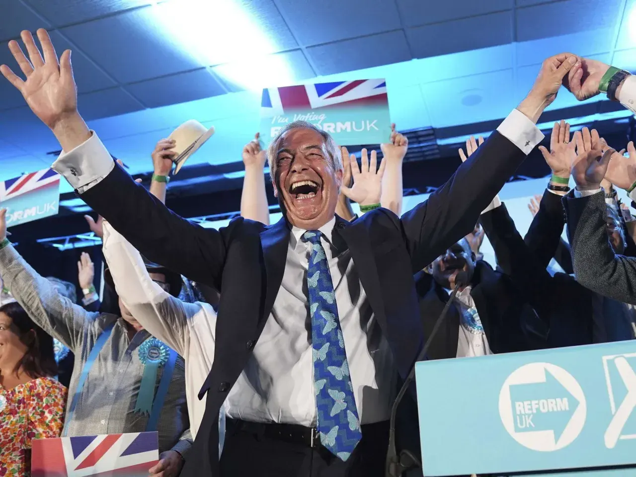 Jubelnde Menge bei einer Wahl in England. Ein Mann mit erhobenen Armen und einem breiten Lachen im Vordergrund, umgeben von Union-Jack-Flaggen und "Reform UK"-Schildern.