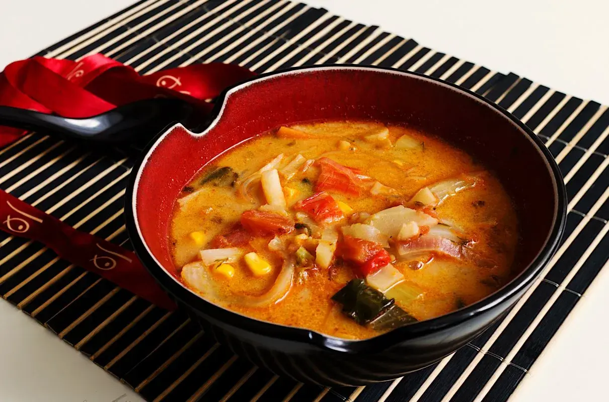 Aromatyczna zupa z surimi z warzywami, podana w misce na macie bambusowej. Obok czarna łyżka i pałeczki ozdobione czerwoną wstążką.