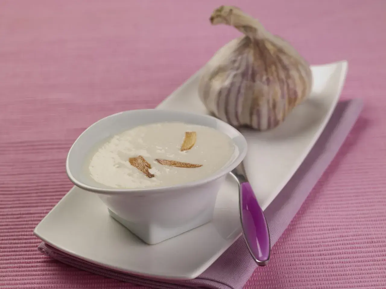 Un bol de crème d'ail onctueuse, garnie de fines lamelles d'ail croustillantes, à côté d'une tête d'ail fraîche.