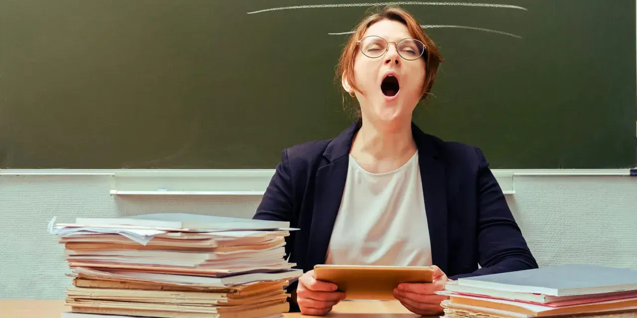 Lehrerin mit Brille gähnt erschöpft hinter einem Stapel Bücher. Ein Klischee, das viele kennen.