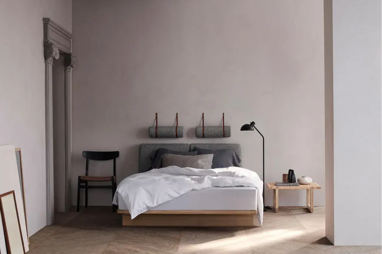 Ein minimalistisches schlafzimmer skandinavisch mit einem Holzbett, grauen Kissen und einer Stehlampe. Zwei gerollte Decken hängen an der Wand.