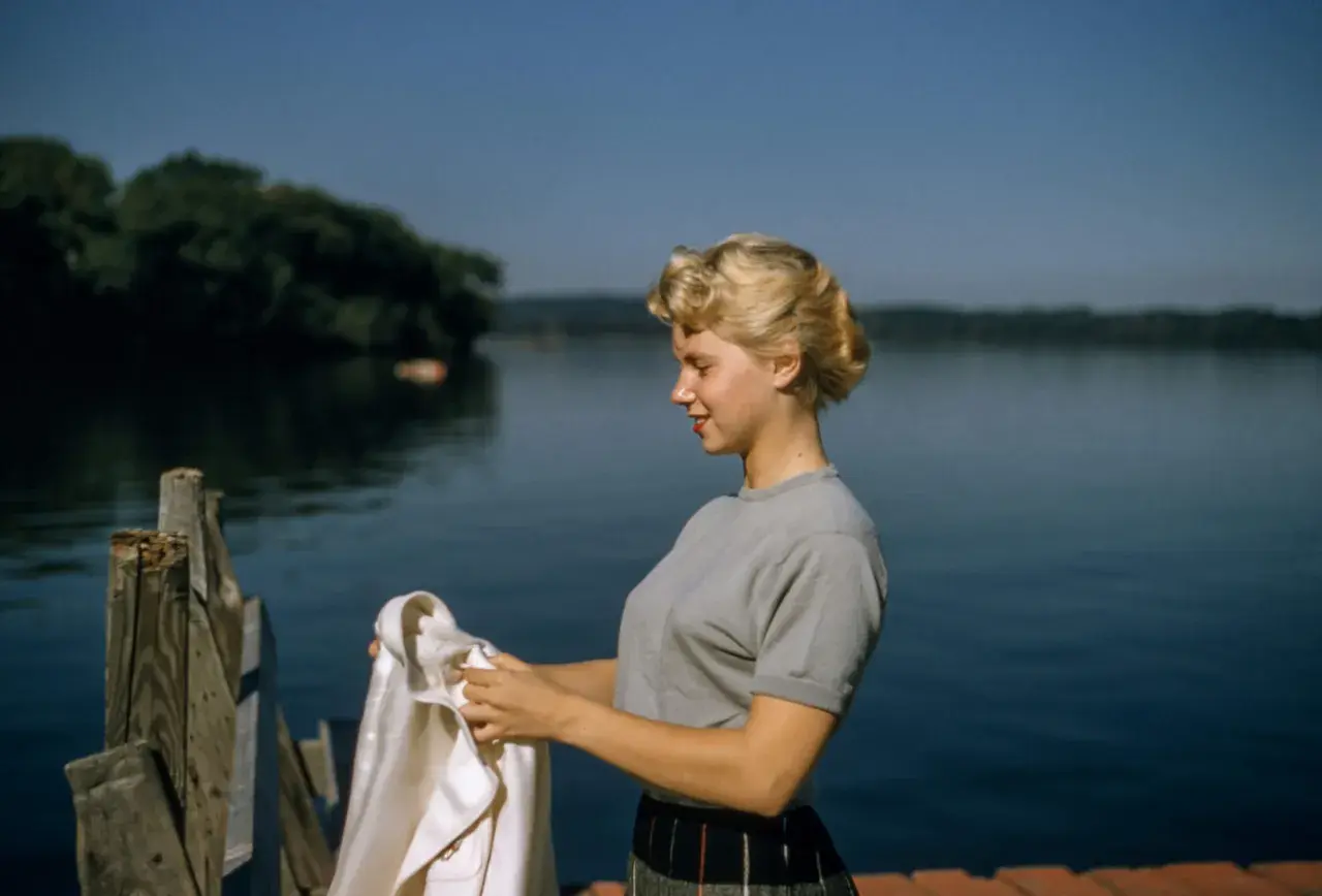 Joven con peinados años 50, con un suéter gris y falda a cuadros, sostiene una chaqueta blanca junto a un lago.