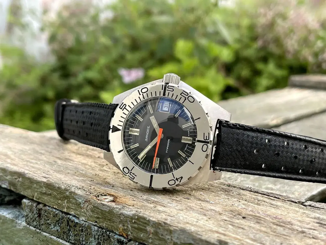 Reloj Sandoz Geneve Typhoon con correa negra, sobre madera rústica y fondo verde.