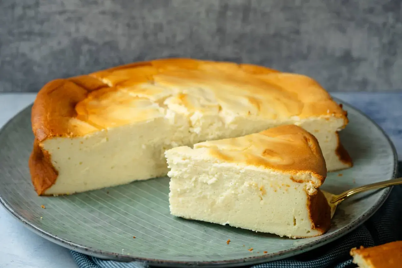 Ein saftiger Quarkkuchen ohne Boden, goldbraun gebacken, liegt auf einem Teller. Ein Stück ist bereits angeschnitten und zeigt die cremige Konsistenz.