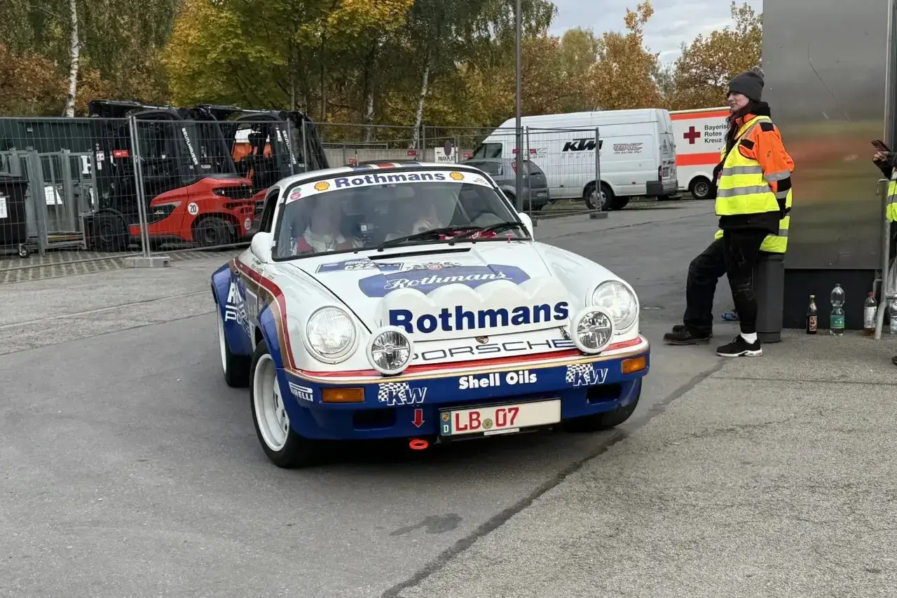 Ein weißer Porsche mit Rothmans-Aufschrift und dem Kennzeichen LB 07. Im Hintergrund stehen Gabelstapler und ein Rettungswagen.