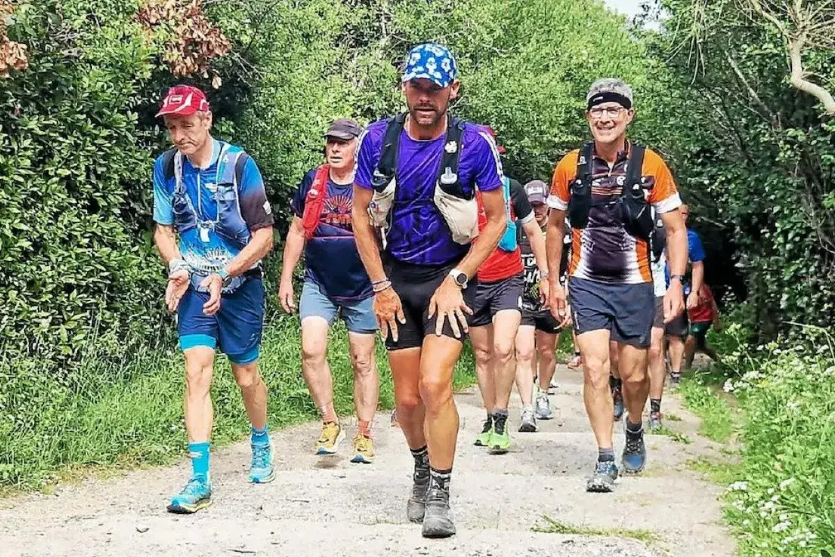 Coureurs en tenue de trail, certains avec des sacs d'hydratation, sur un sentier boisé. Le coureur au centre, portant une casquette bleue, semble en pleine course, peut-être en quête d'un record gr34.