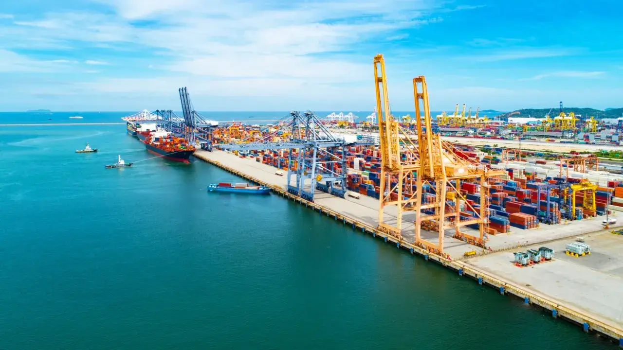 Widok na port kontenerowy z dźwigami, statkami i stosami kontenerów w słoneczny dzień.