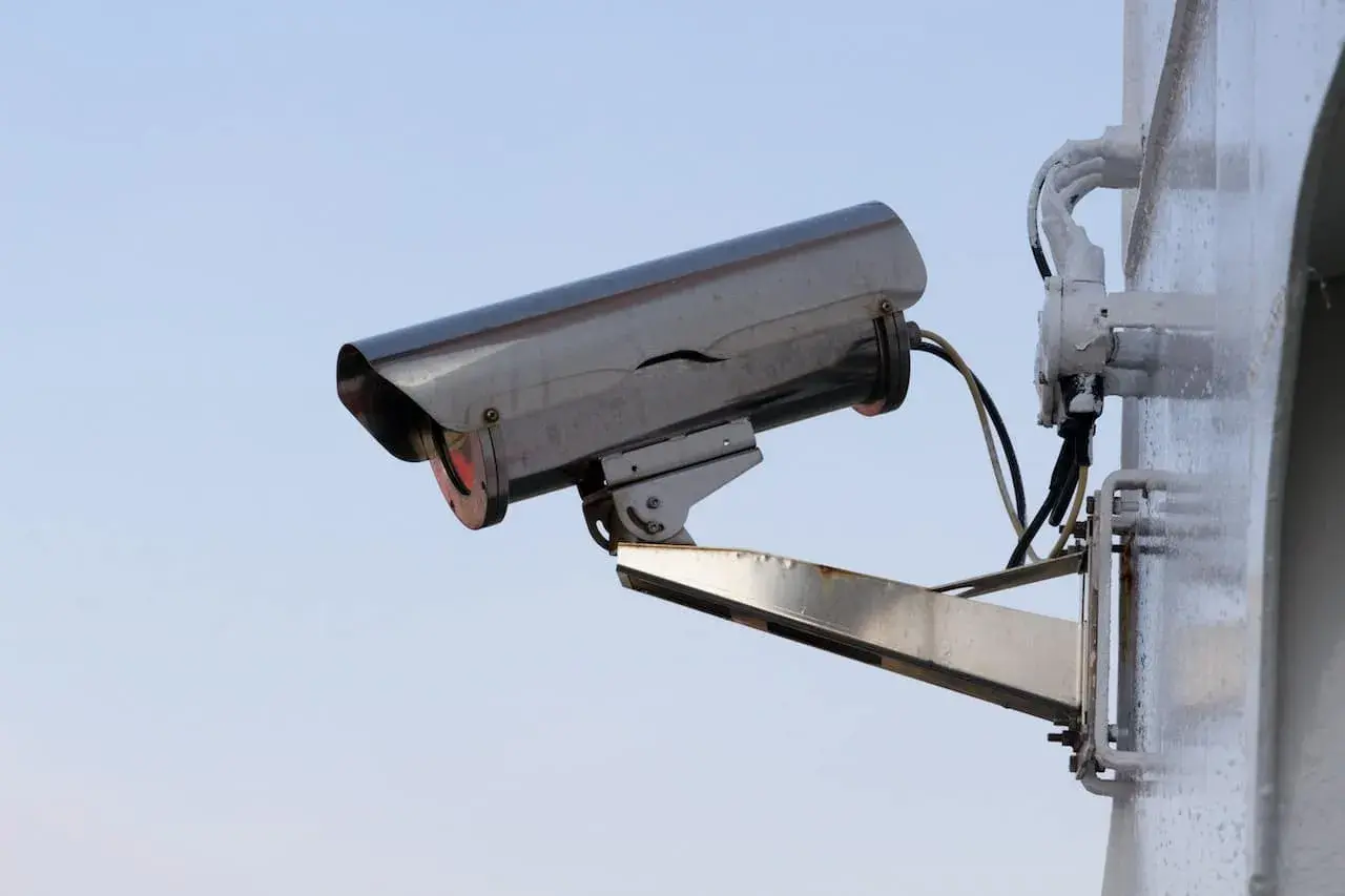 Caméra de surveillance extérieure, fixée sur un mur blanc. Le ciel bleu clair est visible derrière. On dirait qu'il y a un probleme camera de surveillance pas d'image.