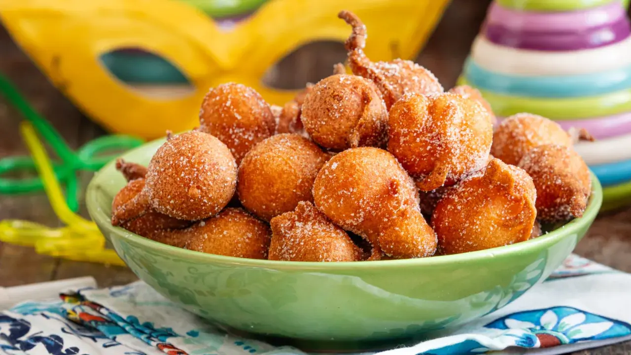 Frittelle di una volta, dolcetti dorati cosparsi di zucchero, pronti per il Carnevale. Maschere e coriandoli sullo sfondo.