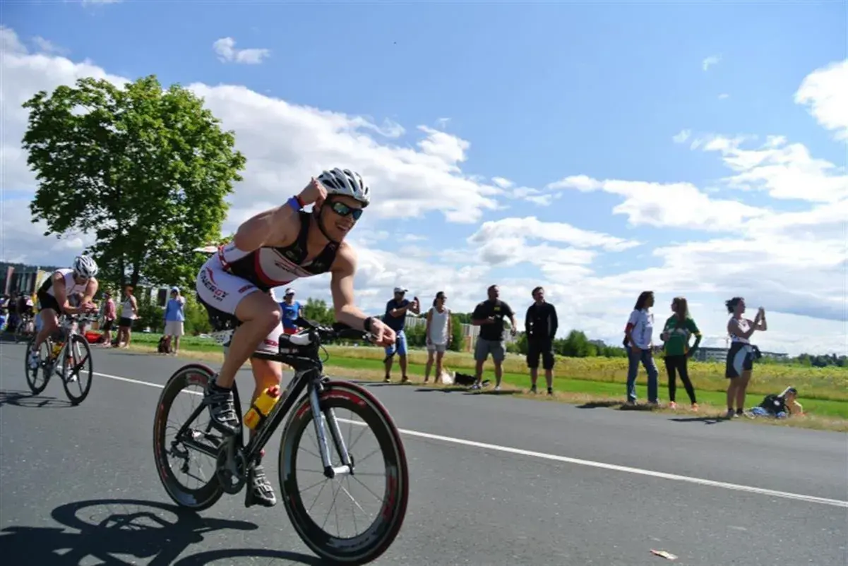 Triathlonista w kasku i okularach ściga się na rowerze, a widzowie dopingują go na poboczu. To część kalendarza triathlonowego.
