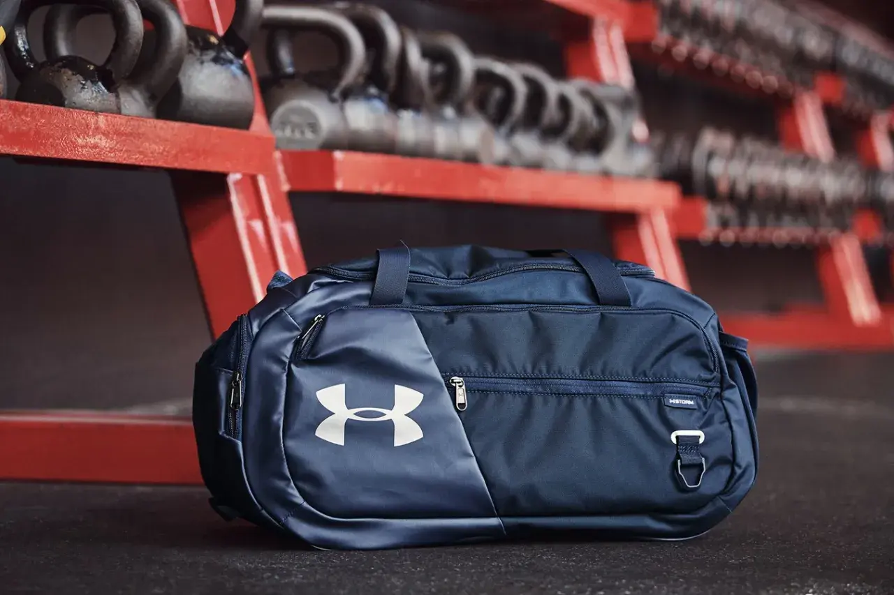 Granatowa torba sportowa 100l z logo Under Armour, gotowa na trening. W tle sztangi i hantle.