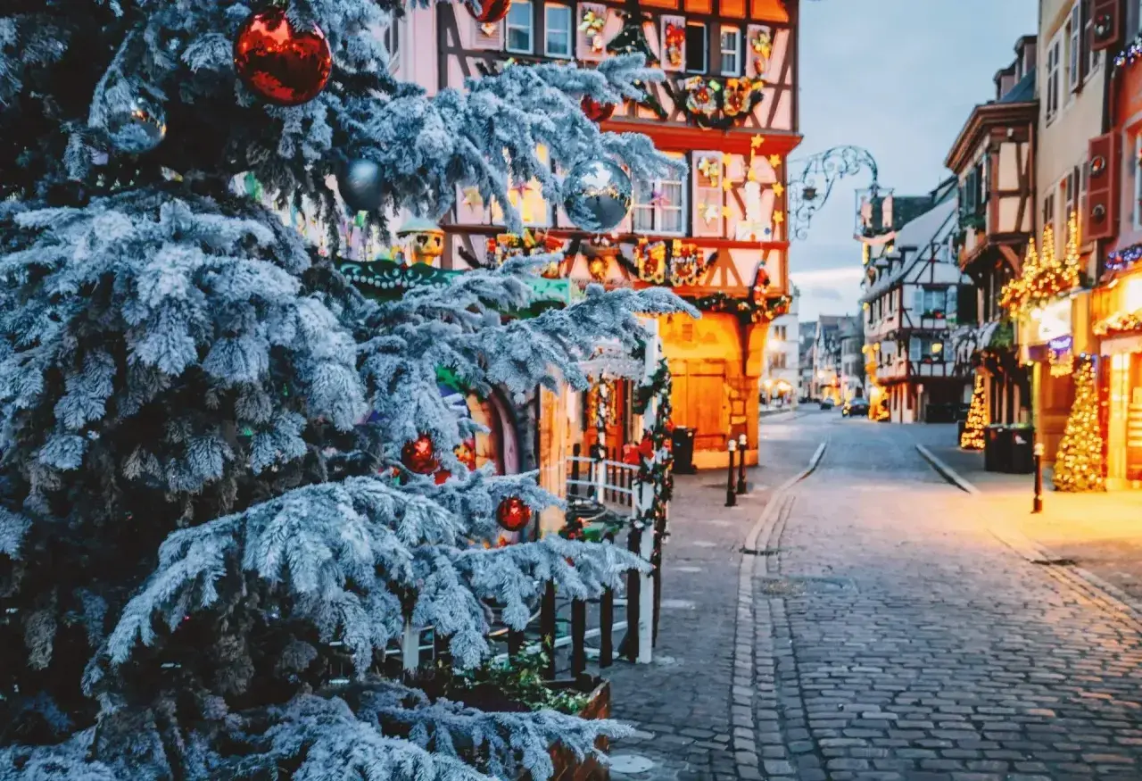 Verschneiter Weihnachtsbaum mit roten Kugeln vor Fachwerkhäusern, die mit Lichtern geschmückt sind. Perfekte reiseziele winter.