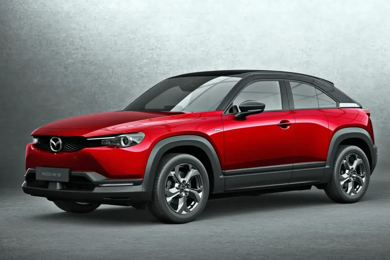 Czerwona Mazda MX-30 z czarnym dachem, stojąca na tle szarej ściany. Stylowy crossover miejski.