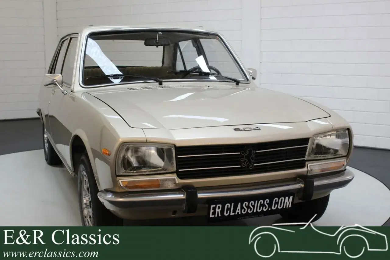 En ljusbeige Peugeot 504 står i ett garage. Bilen har en klassisk design med kromade detaljer.