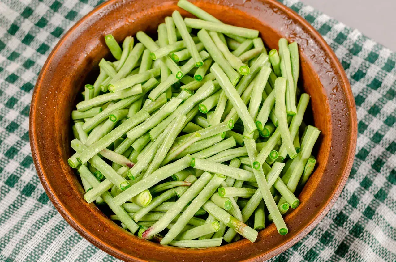 Fagiolini in umido freschi e verdi, pronti per essere cucinati in una ciotola di terracotta su una tovaglia a quadretti.