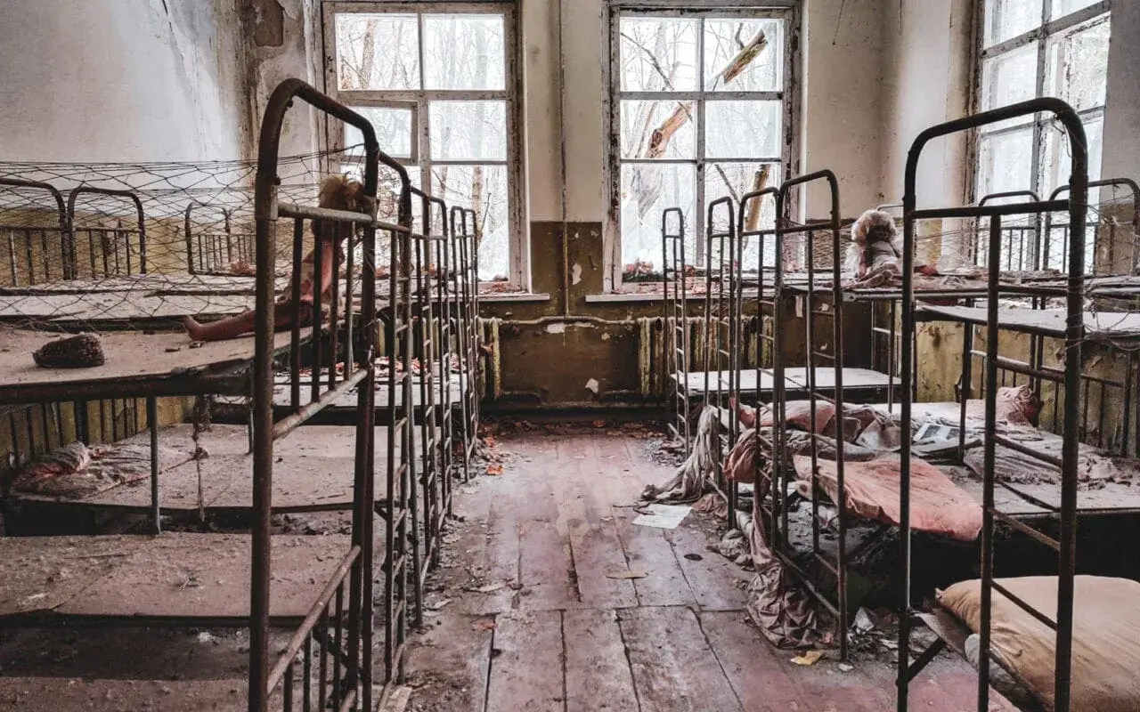 Opuszczony pokój z rzędami zardzewiałych łóżek piętrowych, zniszczonymi oknami i śmieciami na podłodze.