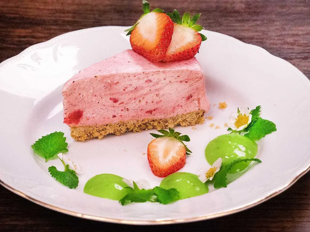 En bit krämig cheesecake jordgubb med färska bär och mynta på en vit tallrik.