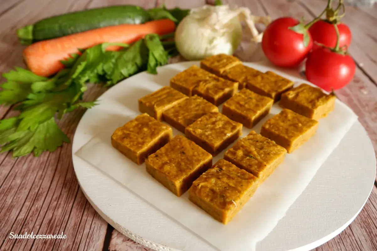 Cubetti di dado vegetale fatto in casa, pronti per insaporire ogni piatto. Verdure fresche come carote, zucchine e pomodori sullo sfondo.