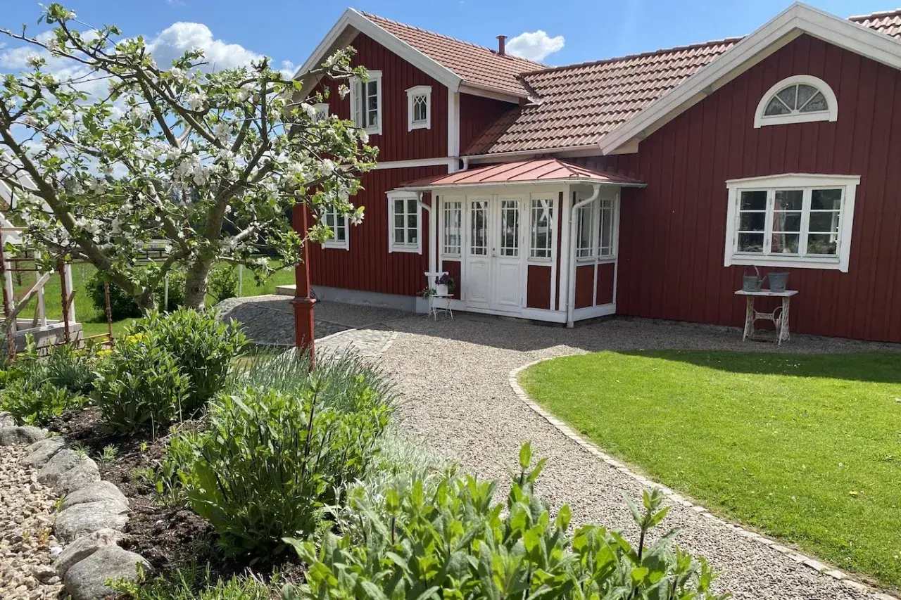 Ett rött hus med vita detaljer och en vacker farstukvist som leder in till hallen. En blommande äppelträd och grönskande trädgård.