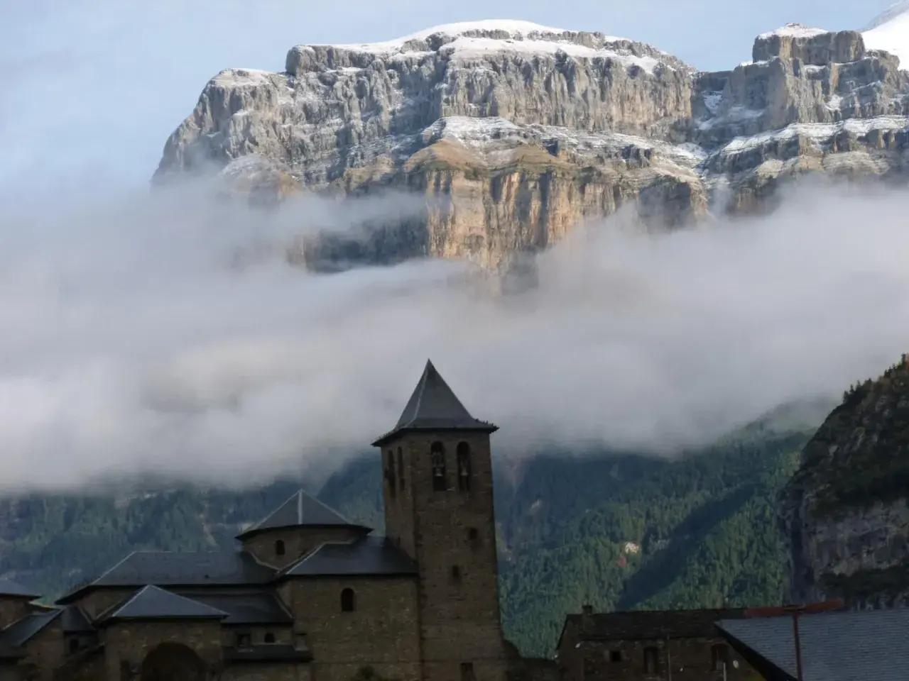 Iglesia románica con campanario, rodeada de niebla, bajo imponentes montañas nevadas. Ideal para explorar rutas desde Torla.