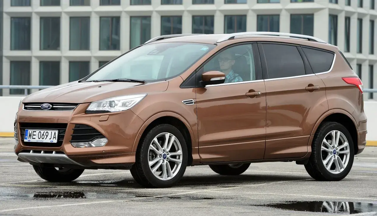 Brązowy Ford Kuga 2 z kierowcą w okularach i kratę, zaparkowany na mokrym parkingu przed nowoczesnym budynkiem.