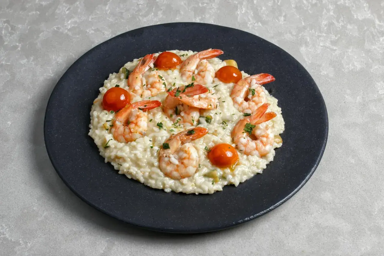 Kremowe risotto z warzywami, ozdobione krewetkami i pomidorkami koktajlowymi, posypane natką pietruszki.
