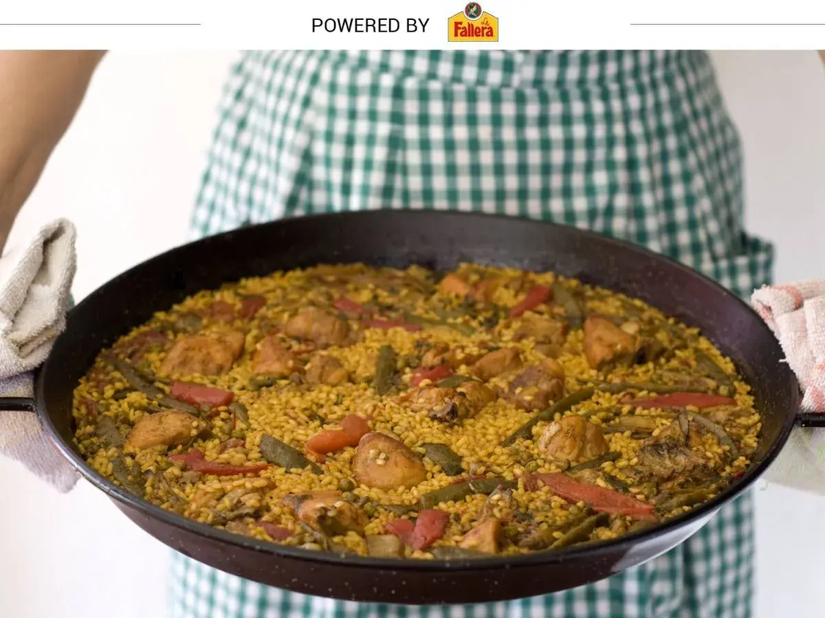 Una persona con un delantal a cuadros verdes sostiene una paella valenciana recién hecha, llena de arroz, pollo, judías verdes y pimientos rojos.