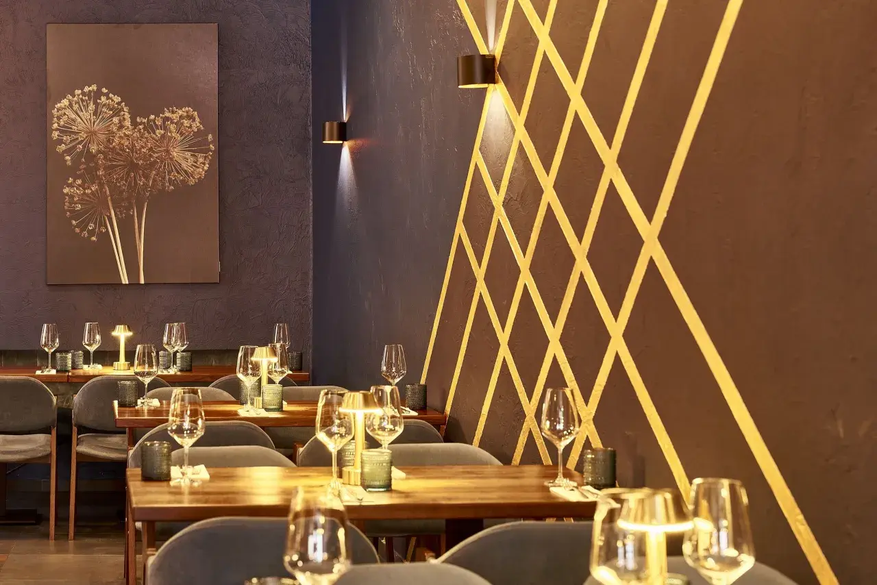 Stilvolles Ambiente im Lovis Restaurant: gedeckte Tische mit Gläsern und Kerzen, dunkle Wände mit goldenem Muster und ein Kunstwerk mit getrockneten Blumen.