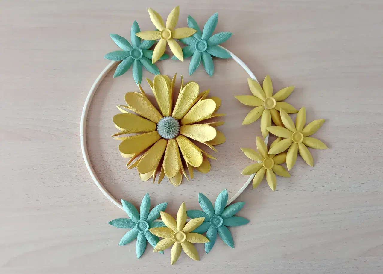 Kreative Blumen aus Eierkarton, gelb und türkis, um einen weißen Ring arrangiert.