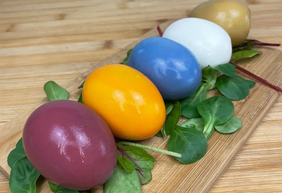 Cinq œufs colorés, du violet à l'orange vif, en passant par le bleu, le blanc et le doré, disposés sur un lit de verdure sur une planche en bois. Chaque couleur oeuf est une surprise.