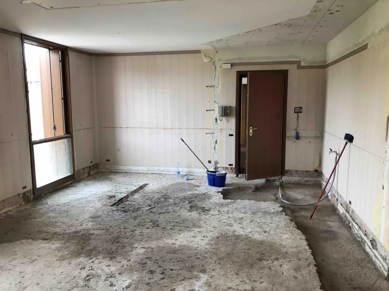 Interno di una stanza in ristrutturazione, con il pavimento in cemento grezzo e pareti spoglie. Si notano attrezzi da lavoro, tra cui un secchio blu e una scopa, che suggeriscono i lavori in corso e i possibili svantaggi dell'intonaco armato.