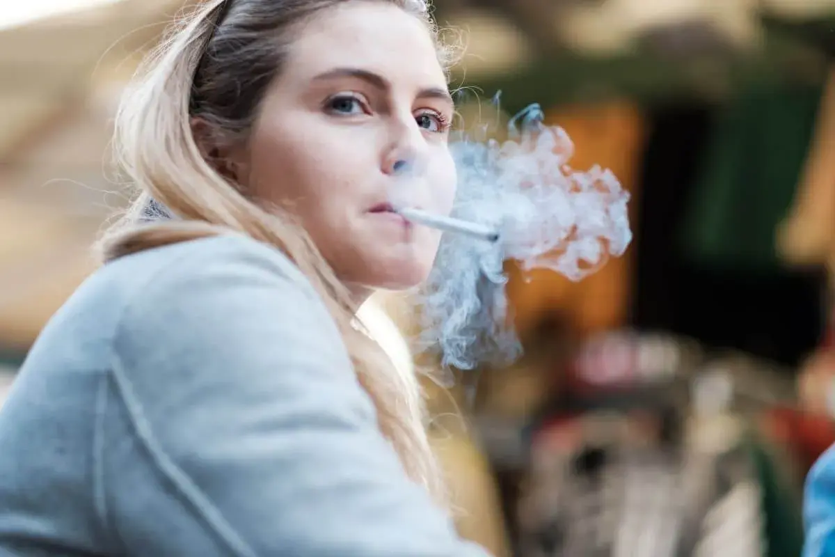 Ragazza bionda con sigaretta tra le labbra, fumo denso che le avvolge il viso. Un momento di pausa, forse dopo essersi lavata i denti, per fumare.