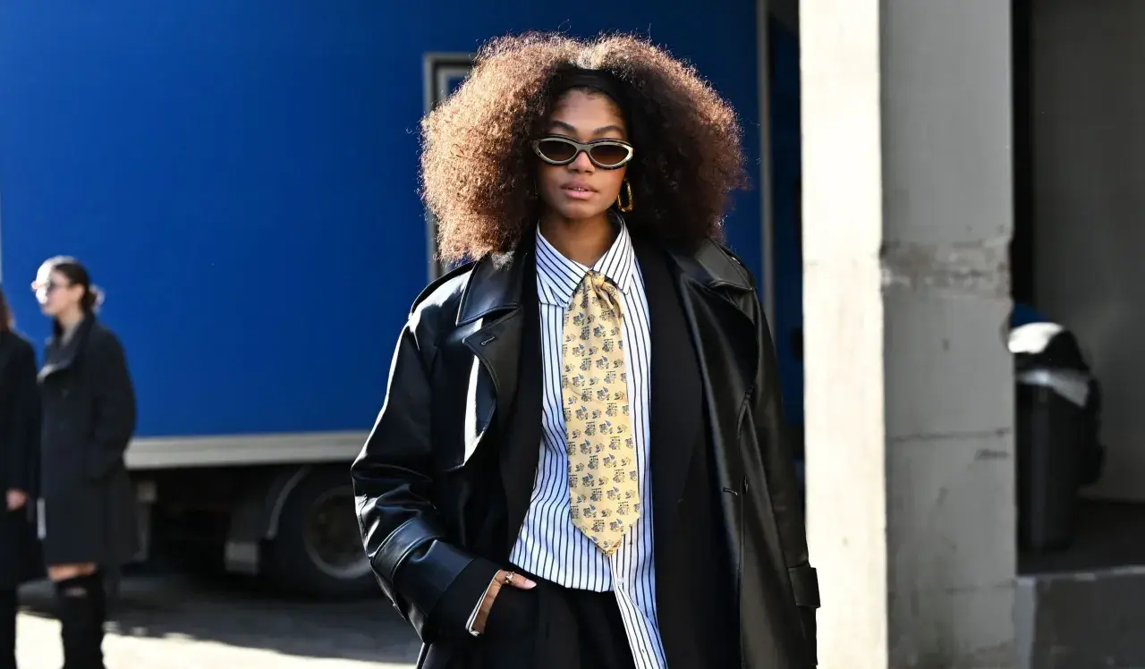 Une femme porte un pantalon noir, une chemise à rayures, une cravate fantaisie et un long manteau en cuir. Ses bottines sont cachées sous son pantalon.