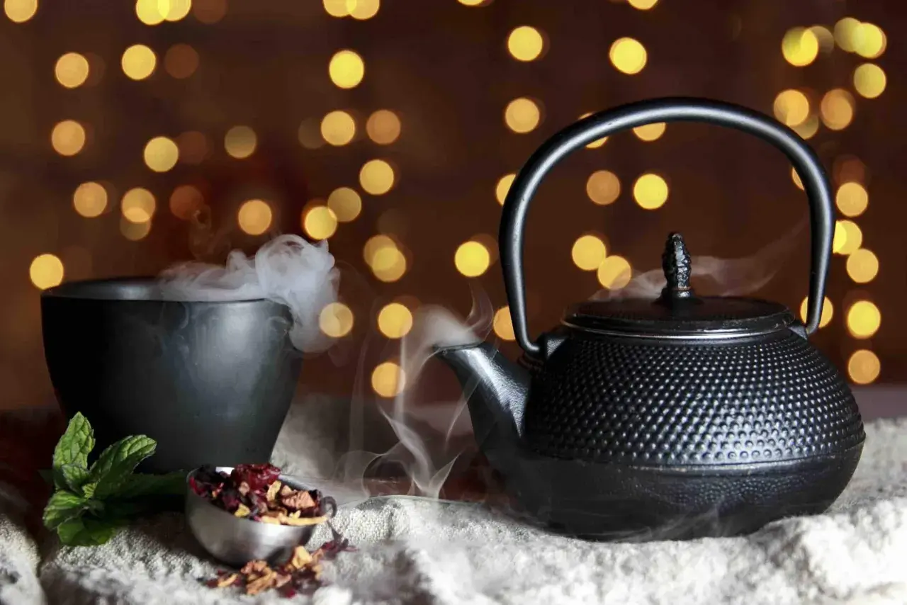 Foto still life: una teiera di ghisa nera emana vapore, accanto a una ciotola fumante e a ingredienti per il tè su uno sfondo di luci soffuse.